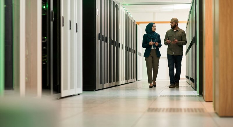 Dua teknisi, seorang wanita berhijab dan seorang pria berpeci, berjalan sambil berbincang di lorong pusat data dengan rak-rak server berlampu hijau lembut.