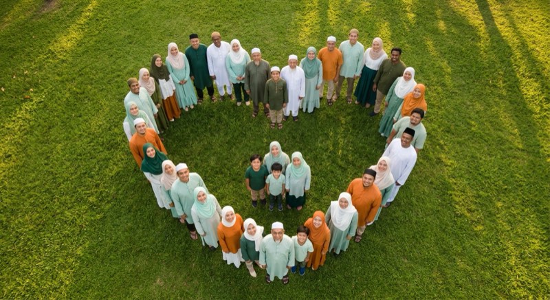 Pemandangan dari atas: umat Muslim dari berbagai usia berpakaian hijau, mint, putih, dan oranye berdiri di rumput membentuk hati.